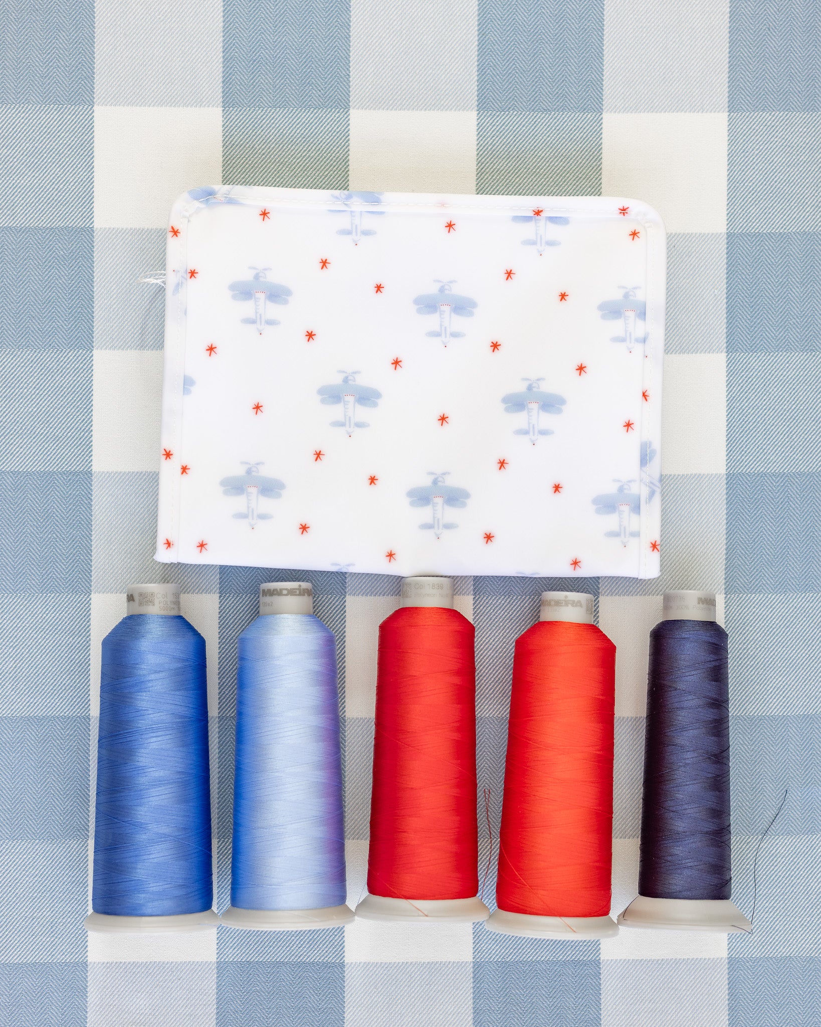 Five spools of thread in blue, red, and purple on a white fabric with airplane pattern against a checkered background.
