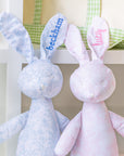 Two plush bunny toys, one blue and one pink, with embroidered names on a white background.