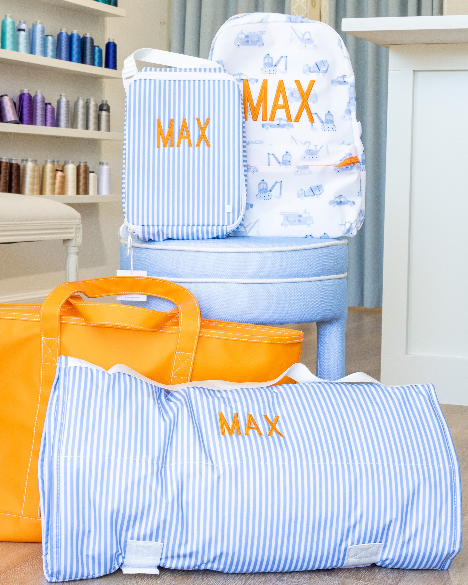 Blue and white striped bags with 'MAX' embroidery, one on a chair, another on the floor, with an orange bag in the foreground.