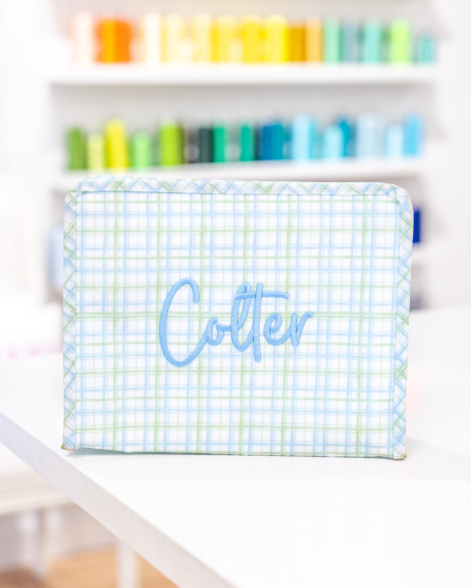 Rectangular container with 'Colter' branding on a checkered pattern, placed on a shelf with colorful items in the background TRVL Classic Plaid Green Large Roadie.