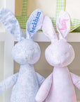 Two plush bunny toys, one blue and one pink, with embroidered names on a white background.